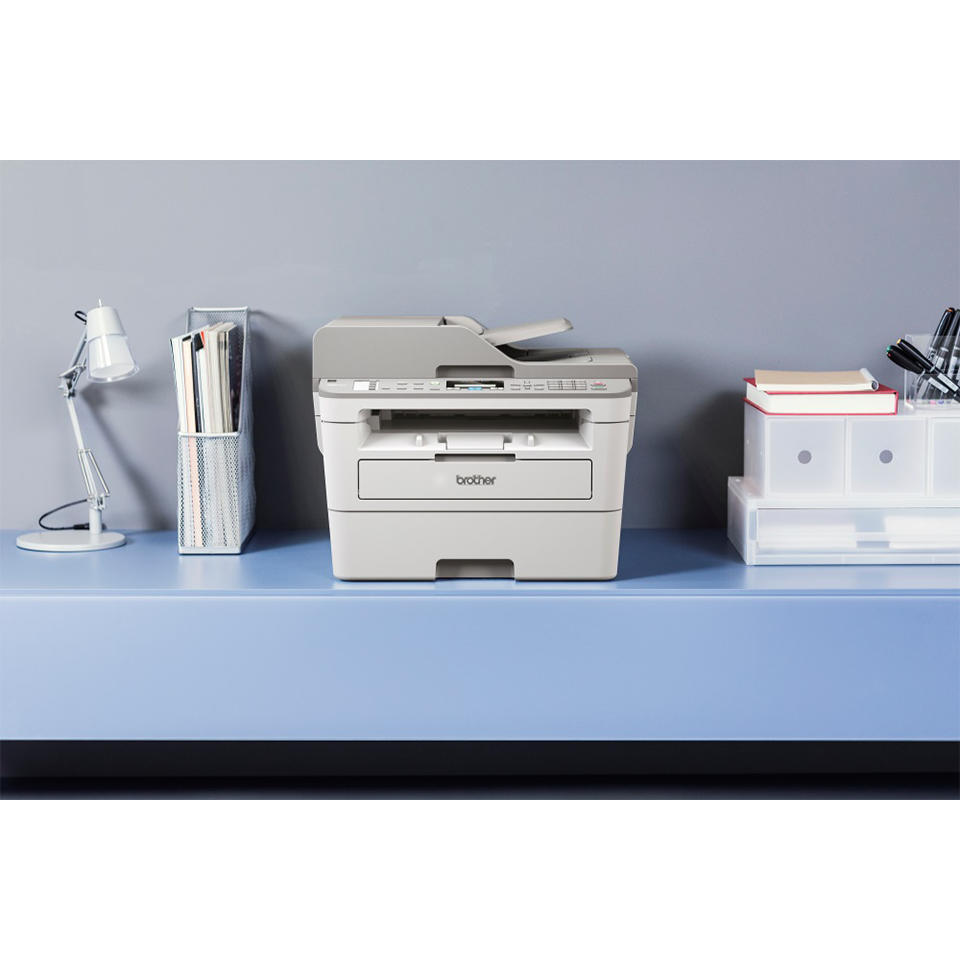 Brother 4-in-1 mono laser MFC-B7715DW printer on a blue desk facing forward