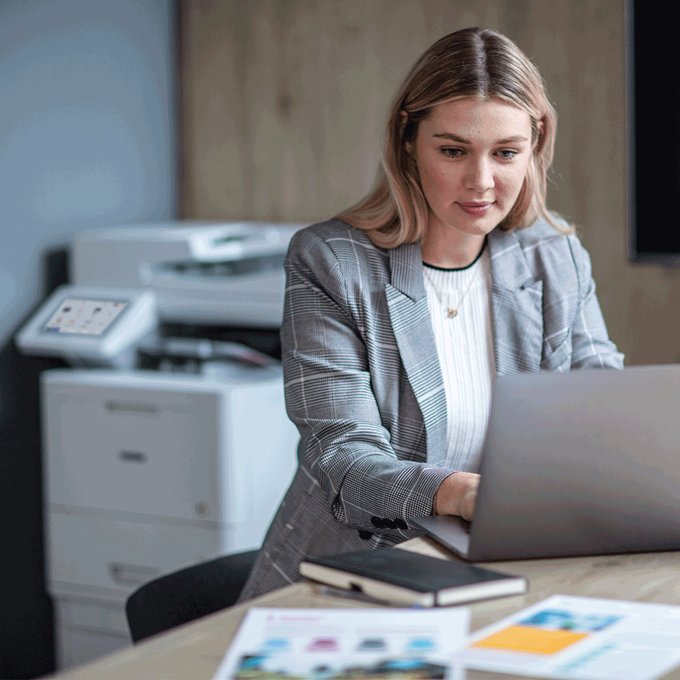 Female sat in office working on laptop, notebook, colour documents, printer