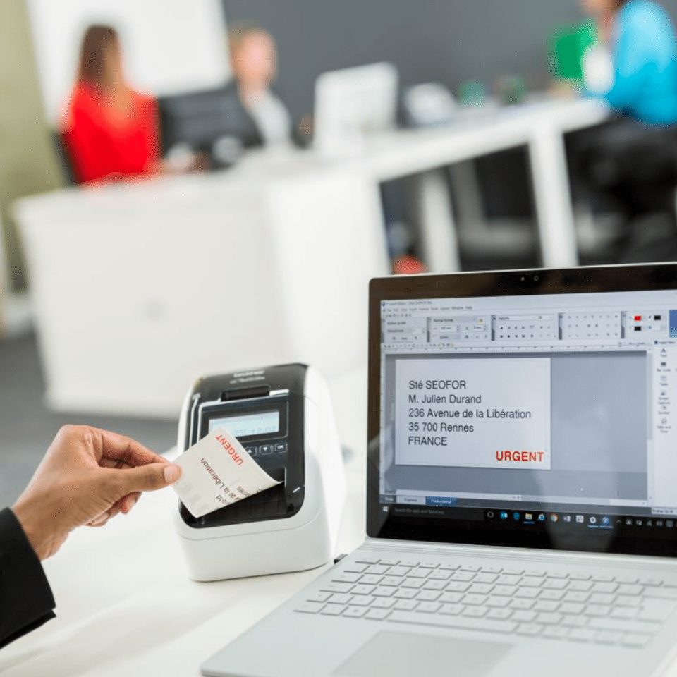 An office worker prints a red and black address or shipping label from their computer using the Brother QL-820NWBVM visitor badge printer