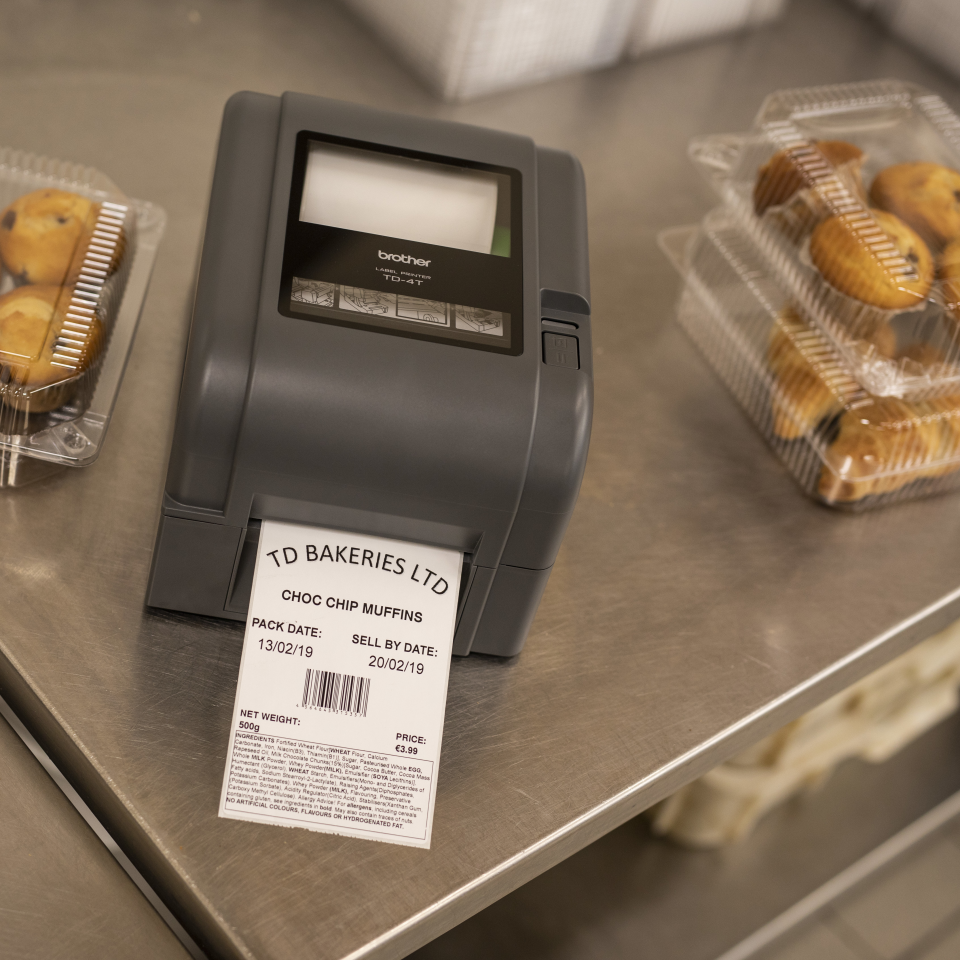 A Brother thermal transfer label printer is on a steel counter next to cakes, with a printed label showing expiry date and allergen ingredients list