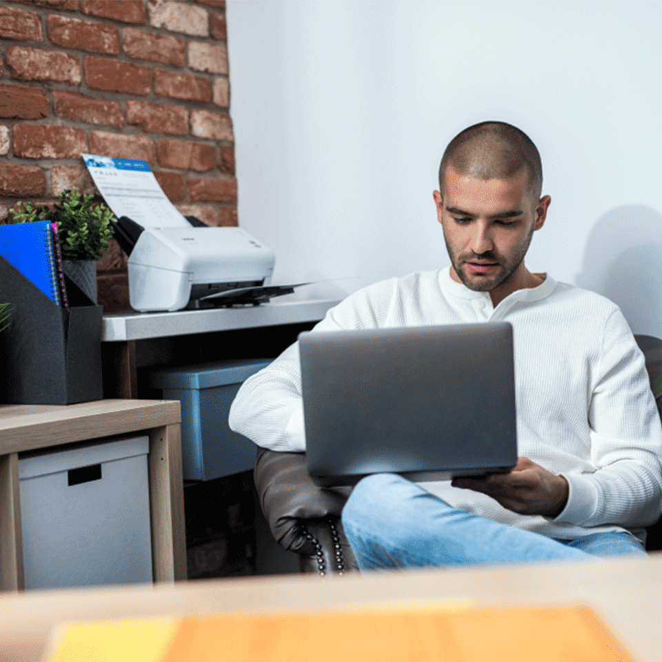Man sat holding laptop, ADS-4100 on desk, plants, folders, notebooks, boxes