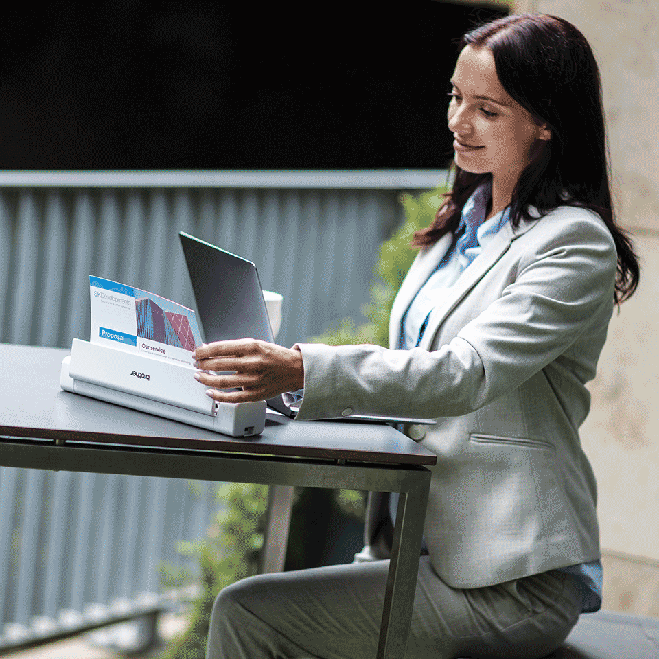Female with brown hair wearing grey suite sat at table using laptop with DSmobile DS-740D portable document scanner