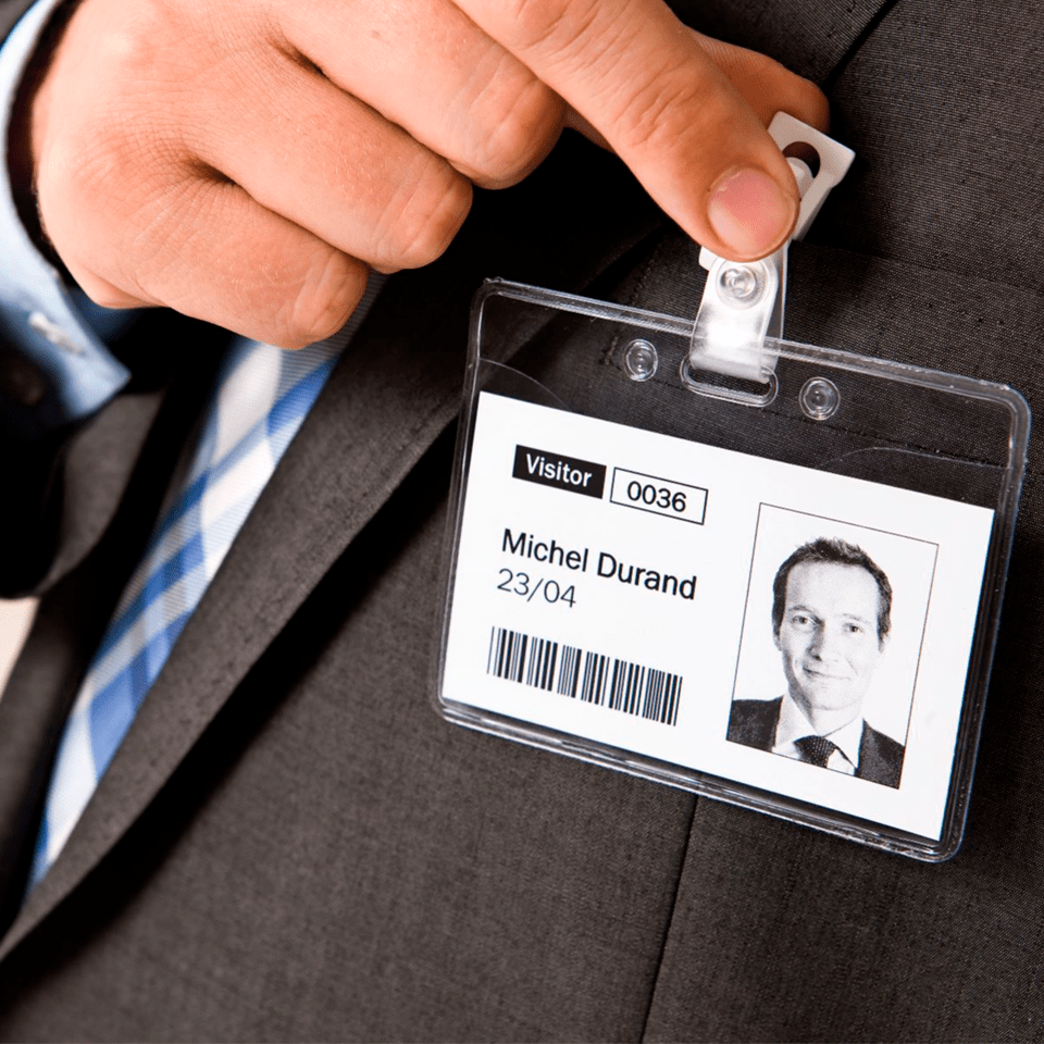 A man clips a visitor badge holder containing a plain paper card visitor identification badge to his grey suit, printed from a Brother QL-820NWBVM visitor and event pass printer