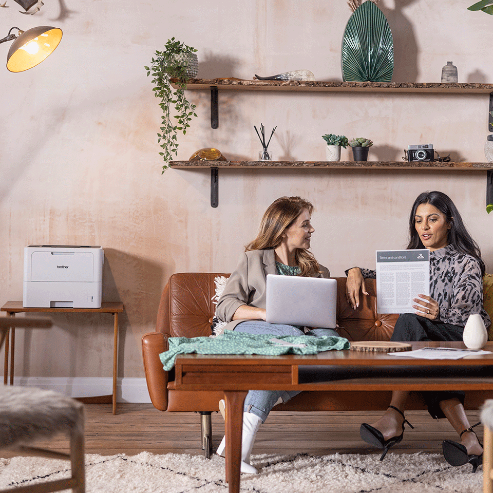 Two females sitting on brown leather sofa with laptop, document, Brother HL-L6210DW printer, shelves