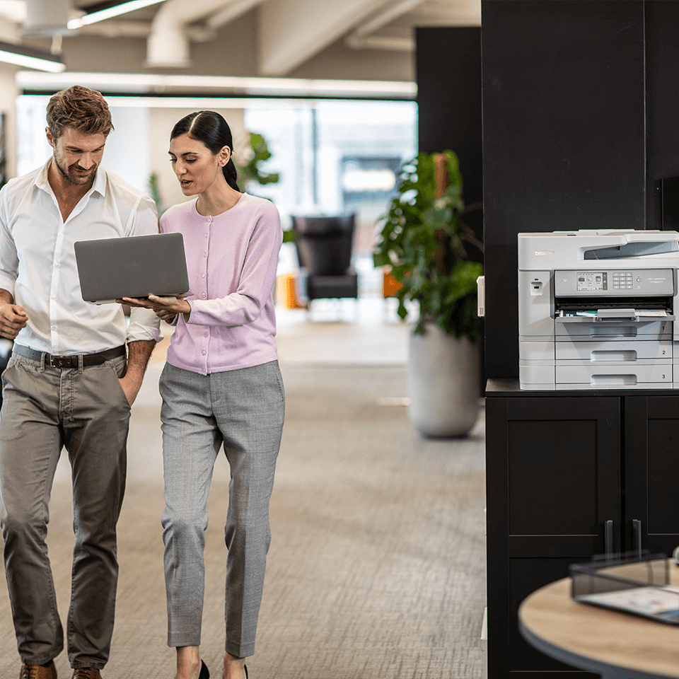 Printer in office, woman holding laptop with ma walking in office, plant, table