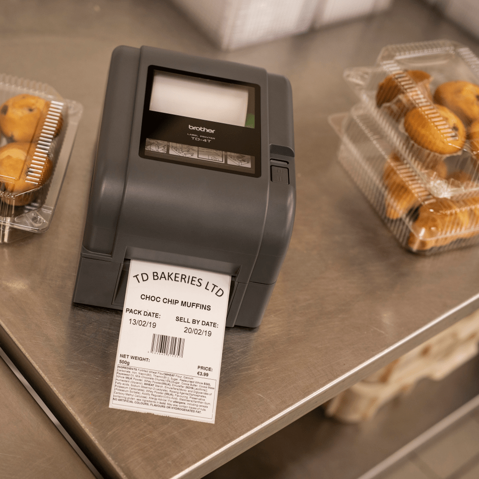 A Brother thermal transfer label printer is on a steel counter next to cakes, with a printed label showing expiry date and allergen ingredients list