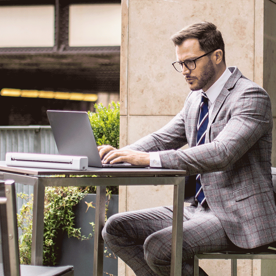 Man wearing grey suit, glasses sat outdoors on table using laptop and DSmobile DS-640 portable scanner
