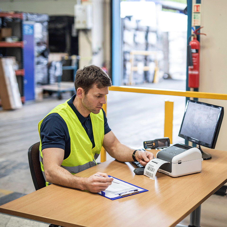 Man wearing hi-vis in warehouse printing labels at desk on computer using Brother TD-4410D desktop label printer