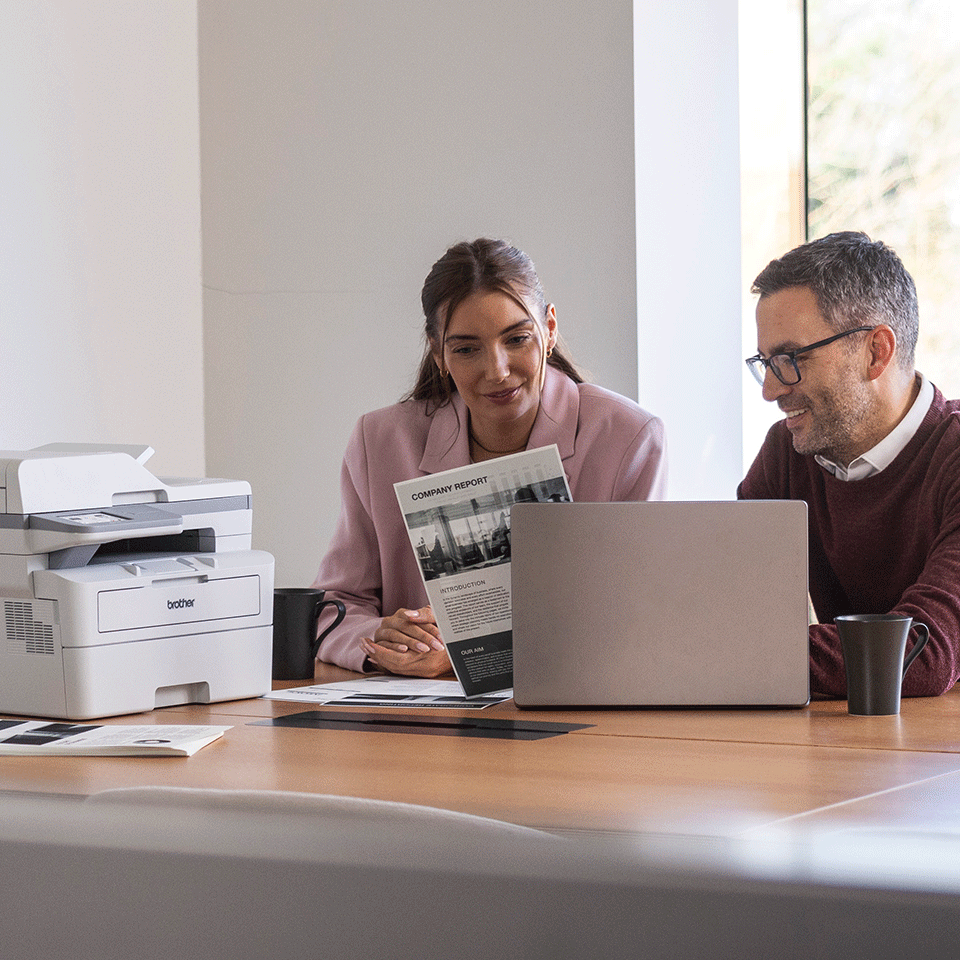 Man and woman sat at desk with Brother MFC-L2960DW, laptop, mug