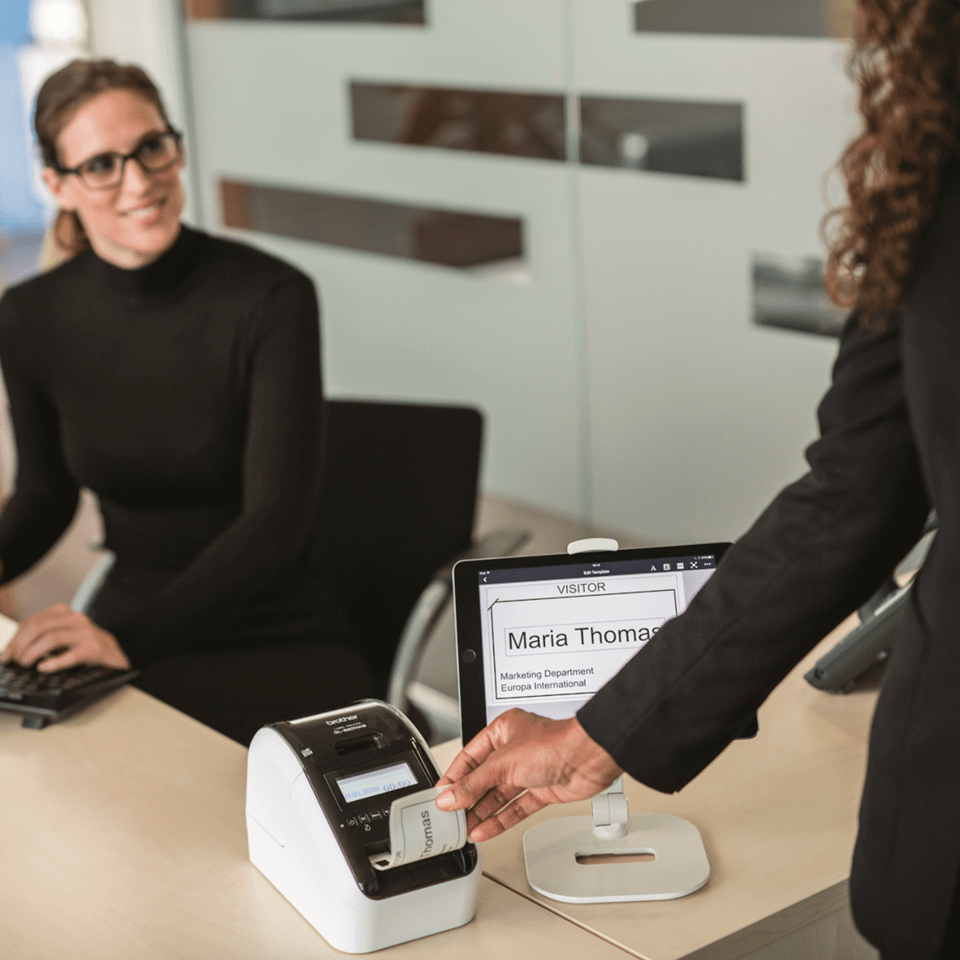 A guest in the reception of a business removes their printed visitor badge from a Brother QL-820NWBVM visitor badge printer after it has been printed using the Brother iPrint&Label app on an iPad