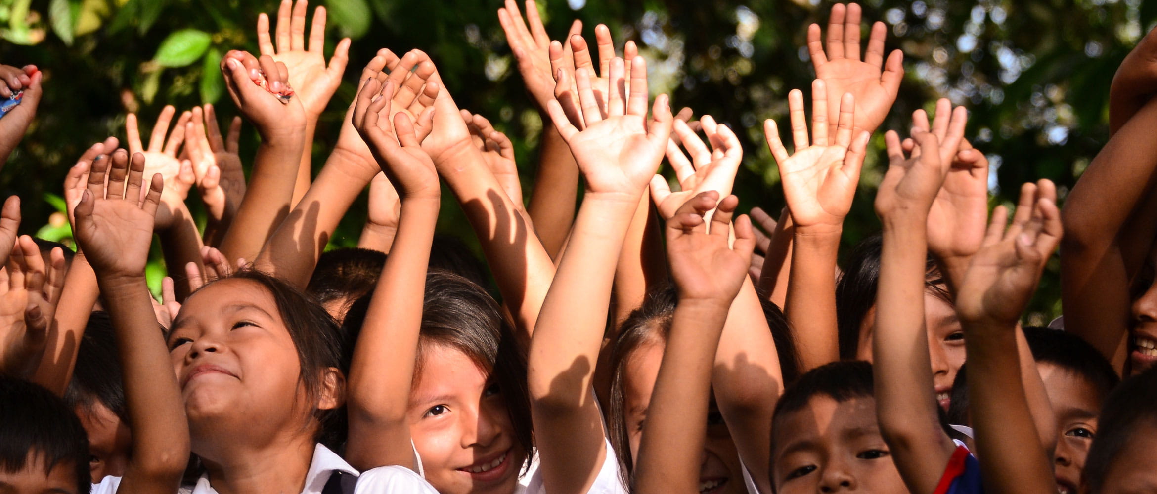 Closeup of a group of children smiling while holding their hands in the air with greenery in the background