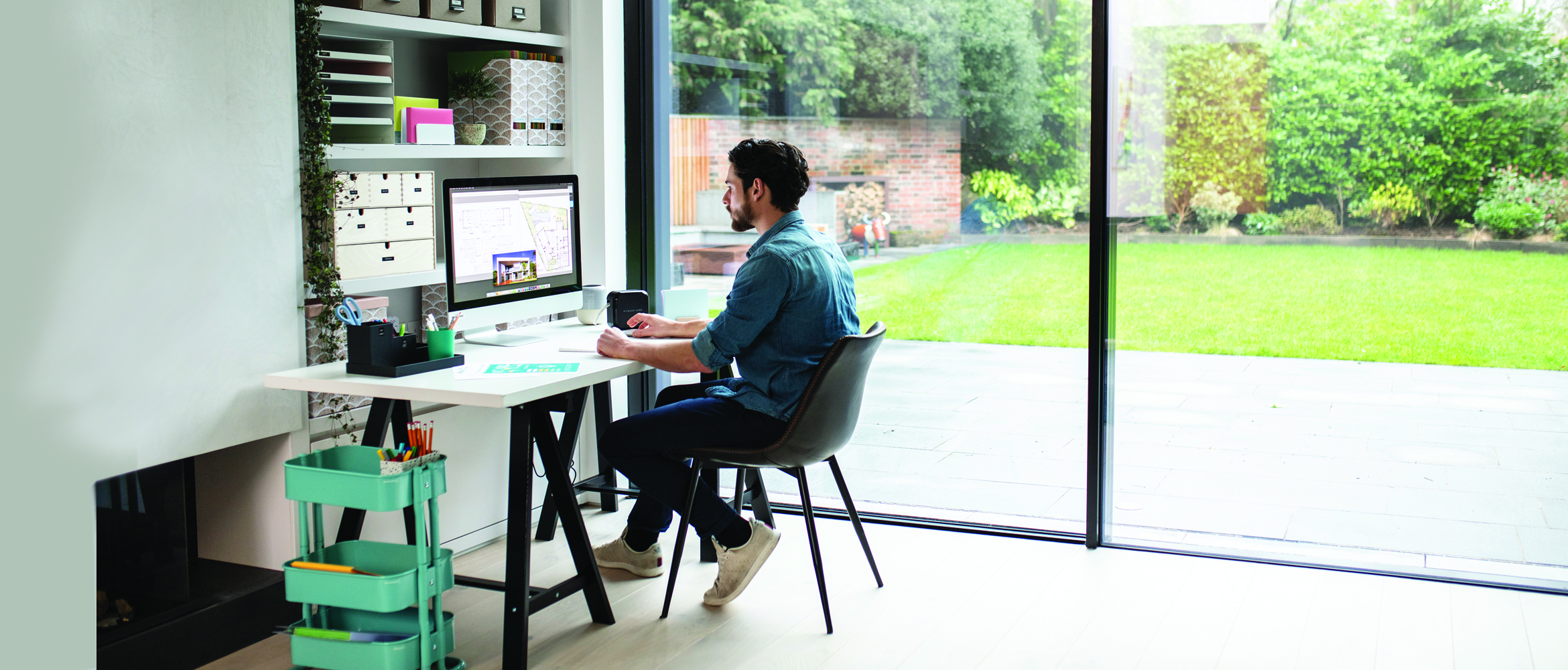 Man sat at desk looking at computer screen with shelves behind and a view of the garden through the patio doors