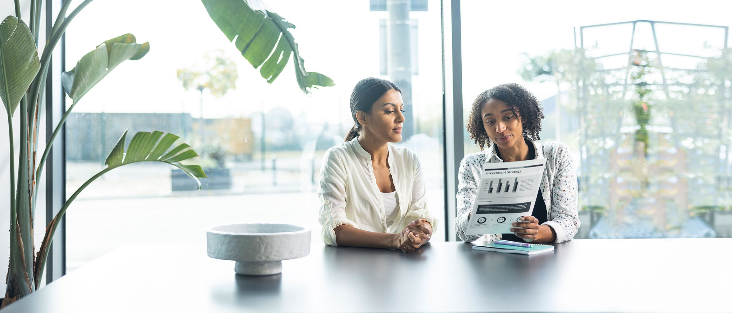 Two women sat at a large grey desk, one holding black and white document, plants, large window