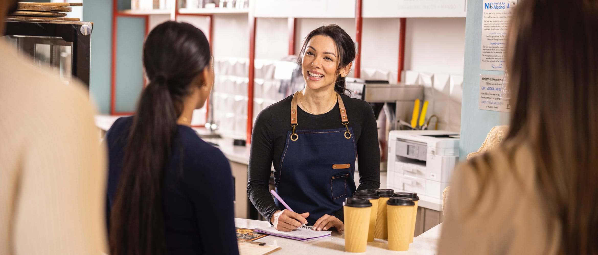 A female retail assistant taking an order from a woman in a busy coffee shop with a printer in the background