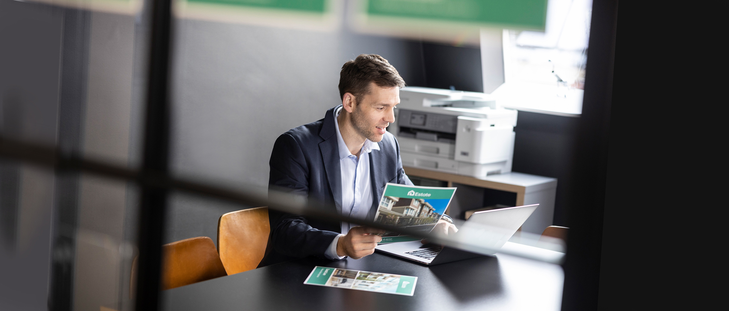 Man sitting at desk holding colour documents, printer, chair, cabinet