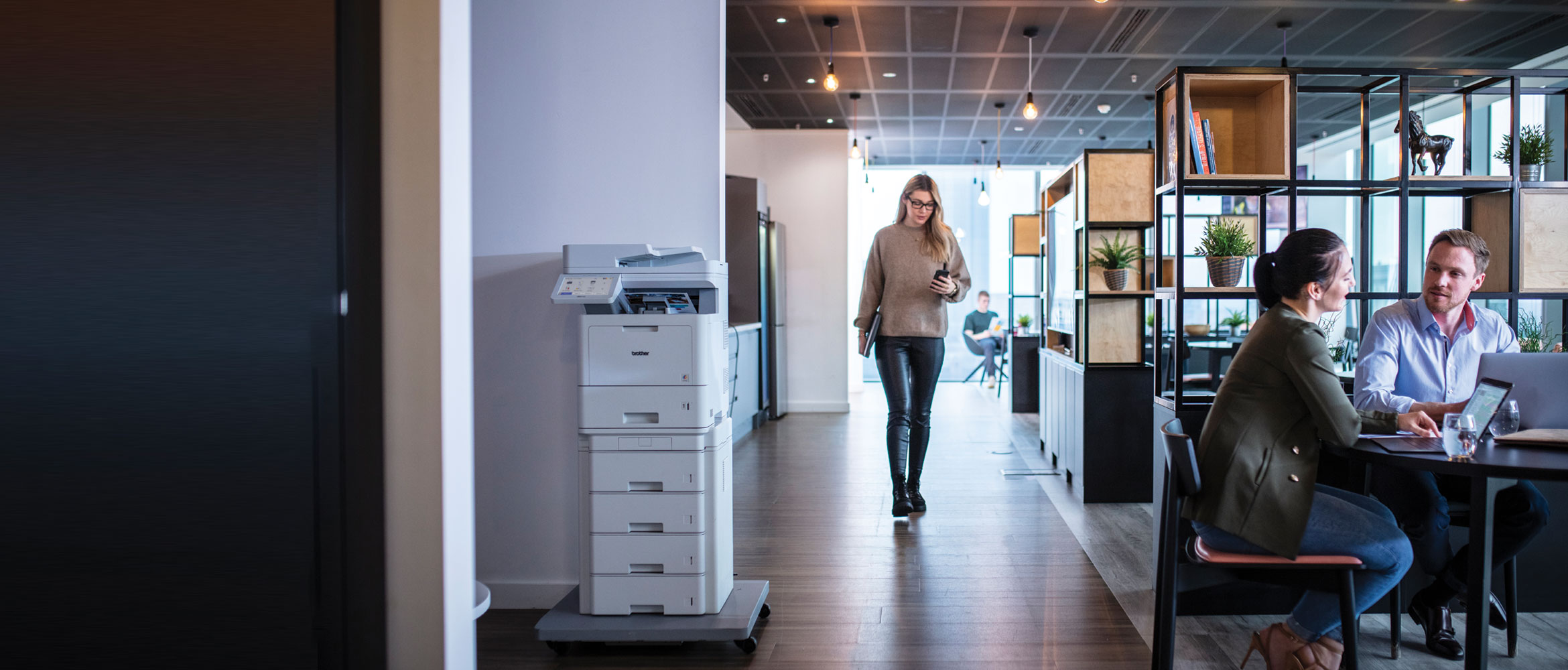 Man and woman sat at a table with a woman walking by and a printer in the corner