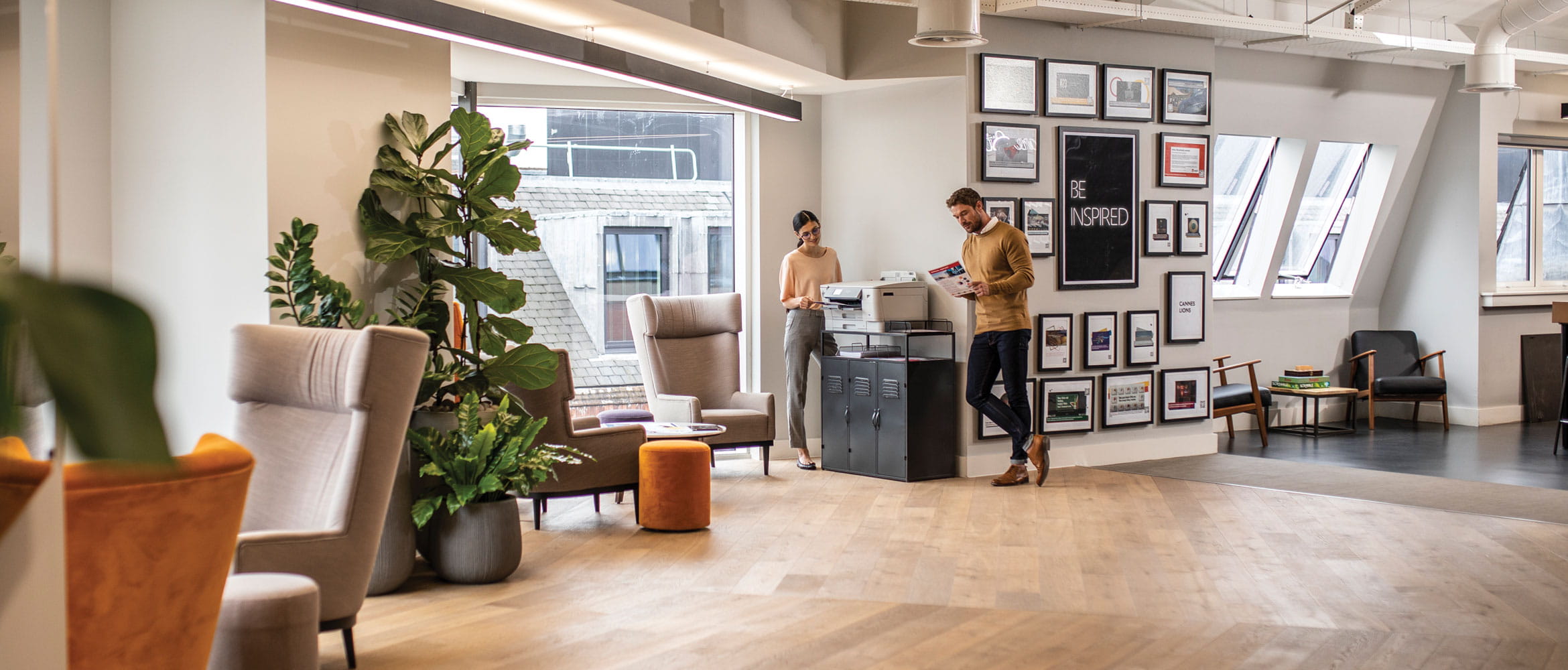 A man and a woman stood next to a printer reading in a large room with windows and chairs