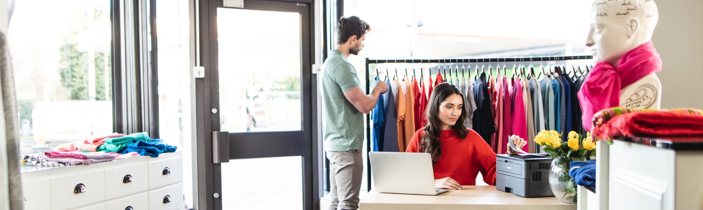 A female sales assistant sat at a desk, printing a document in a clothes shop with a male customer browsing stock in the background