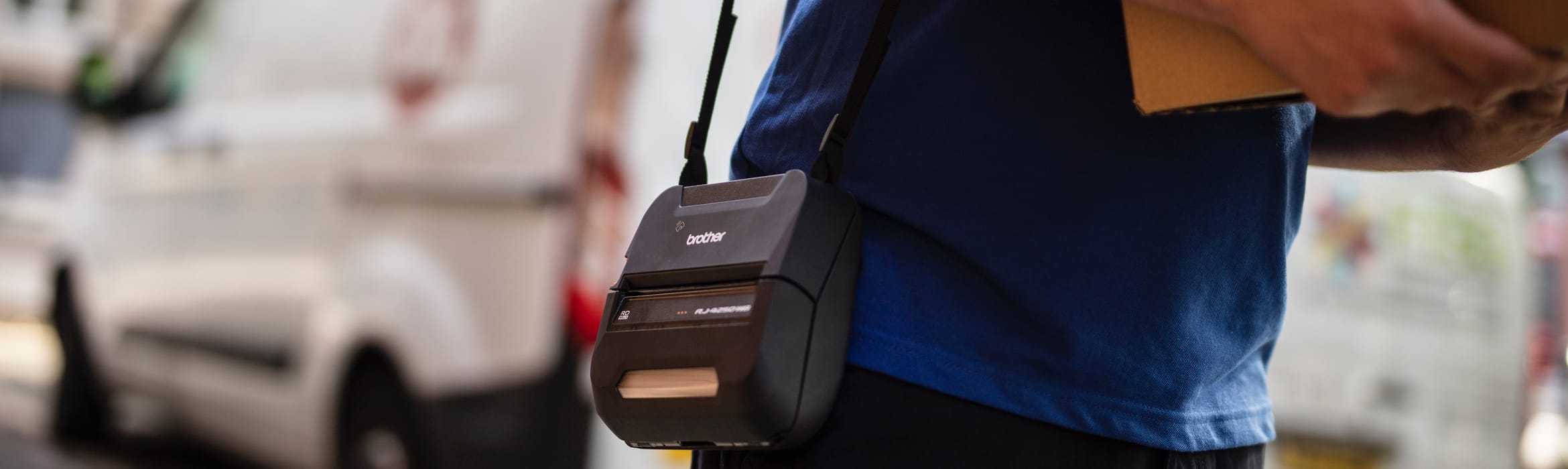 Closeup of a delivery driver carrying a brown cardboard box with a Brother RJ label printer over his shoulder and white vans in the background