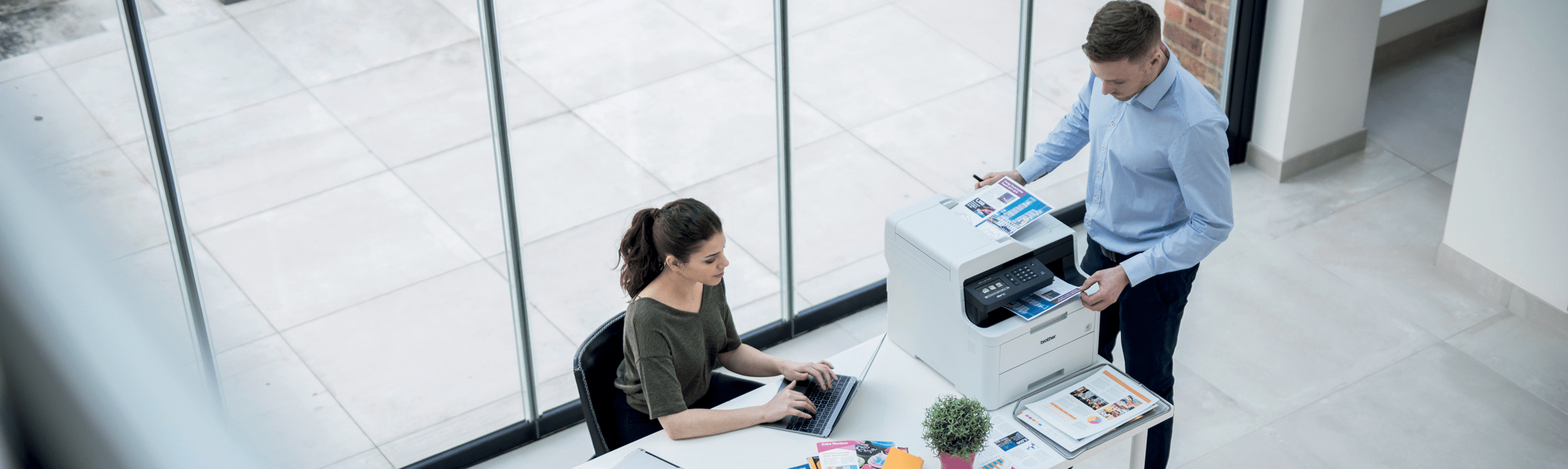 Man sat at desk with laptop and DCP-L3550CDW in background