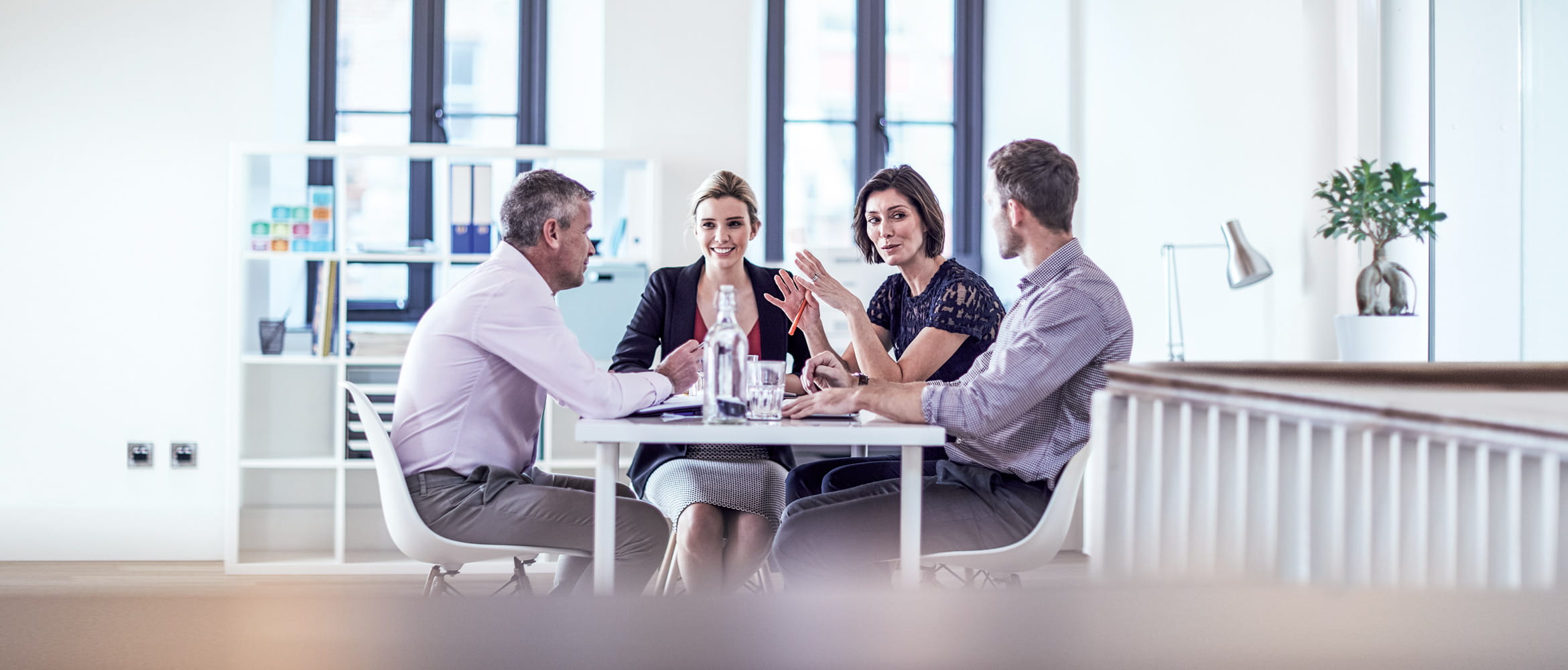 Two men and two woman sat around a table in an office, plant, shelving, windows