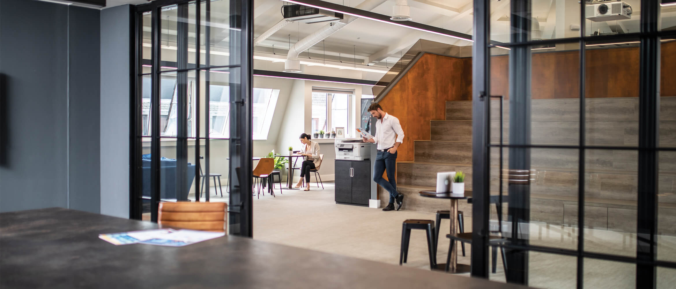 Man in office printing document, woman sat working at desk, meeting room, chair, table, documents