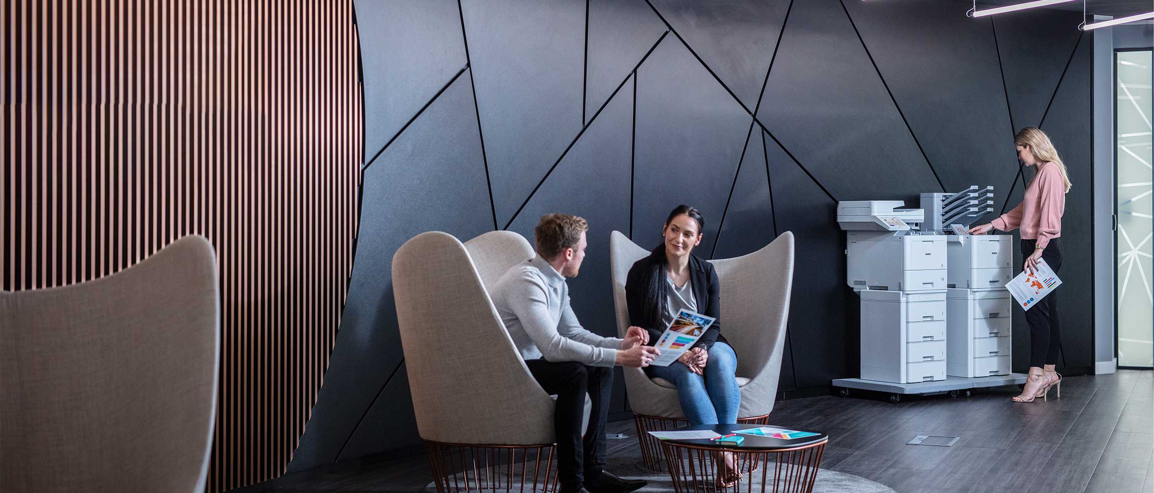 Male and female sat talking around small table, female in background stood next to printers