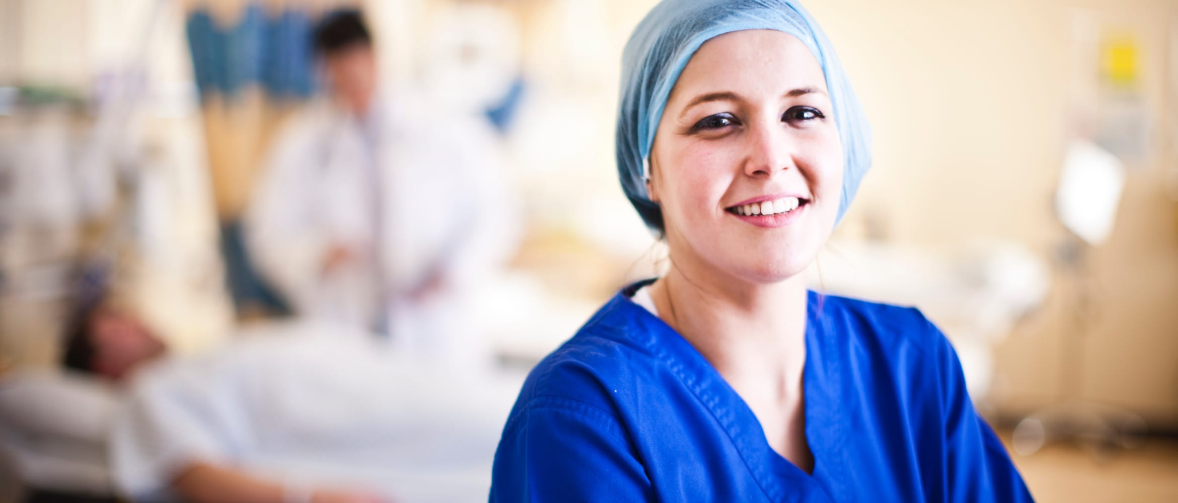 Healthcare professional wearing a blue top and head covering in a hospital ward with a doctor attending a patient in the background