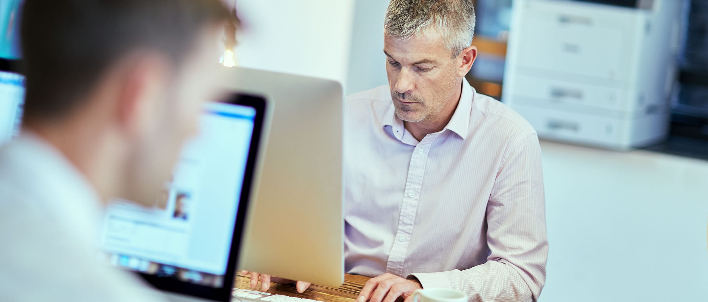 Two men sat at desk working on computers with laser printer in the background