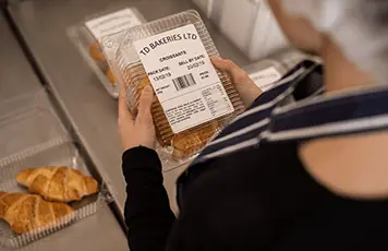 Woman wearing blue and white striped apron holding plastic bakery product with label