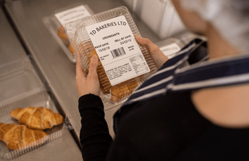 Woman wearing blue and white striped apron holding plastic bakery product with label