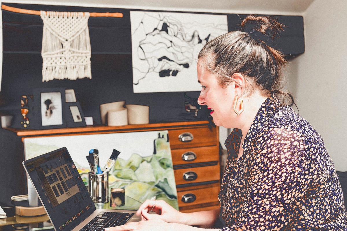 Andrea Ainsworth in her studio, working on a notebook computer while sat at a desk with artwork hanging in the background