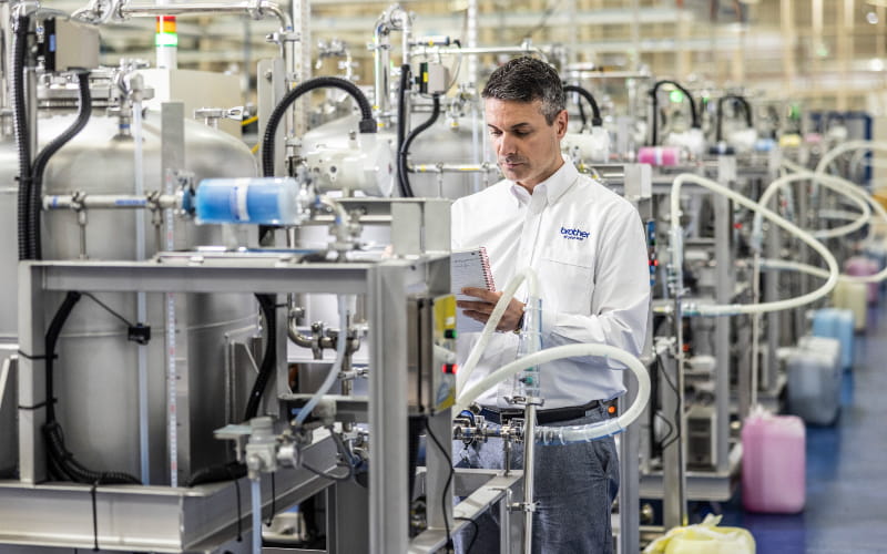 Man holding clipboard in a factory surrounded by machinery, pipes and containers filled with ink