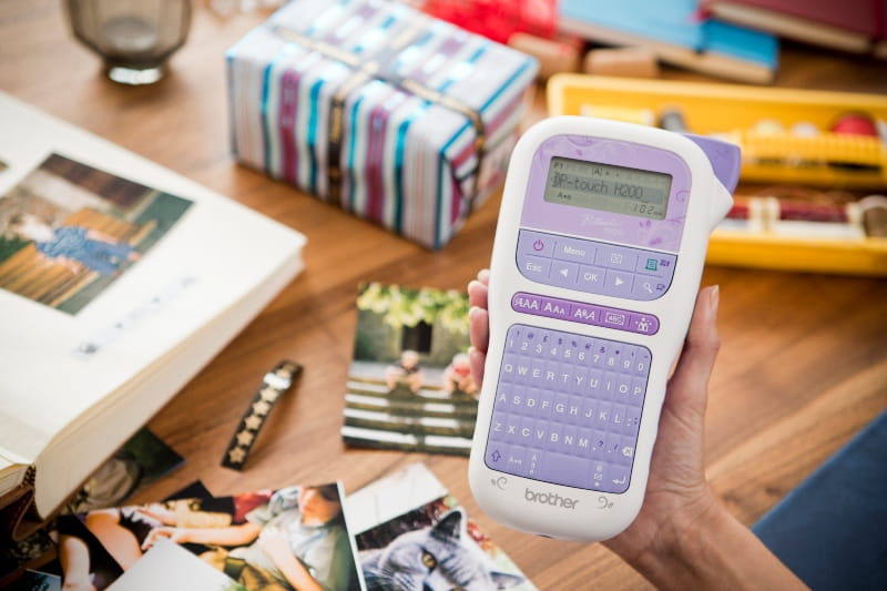 Hand holding purple p-touch labelling device with a table in background with a gift-wrapped box tied with printed ribbon, an open photo album and some photos