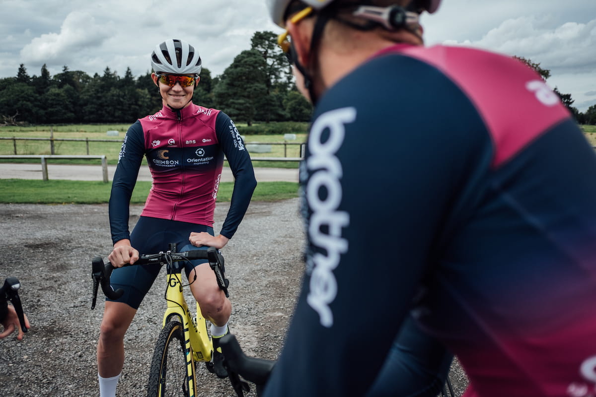 Racing cyclist, purple and blue jersey, blue shorts, white helmet, sat on yellow bike, facing camera, smiling