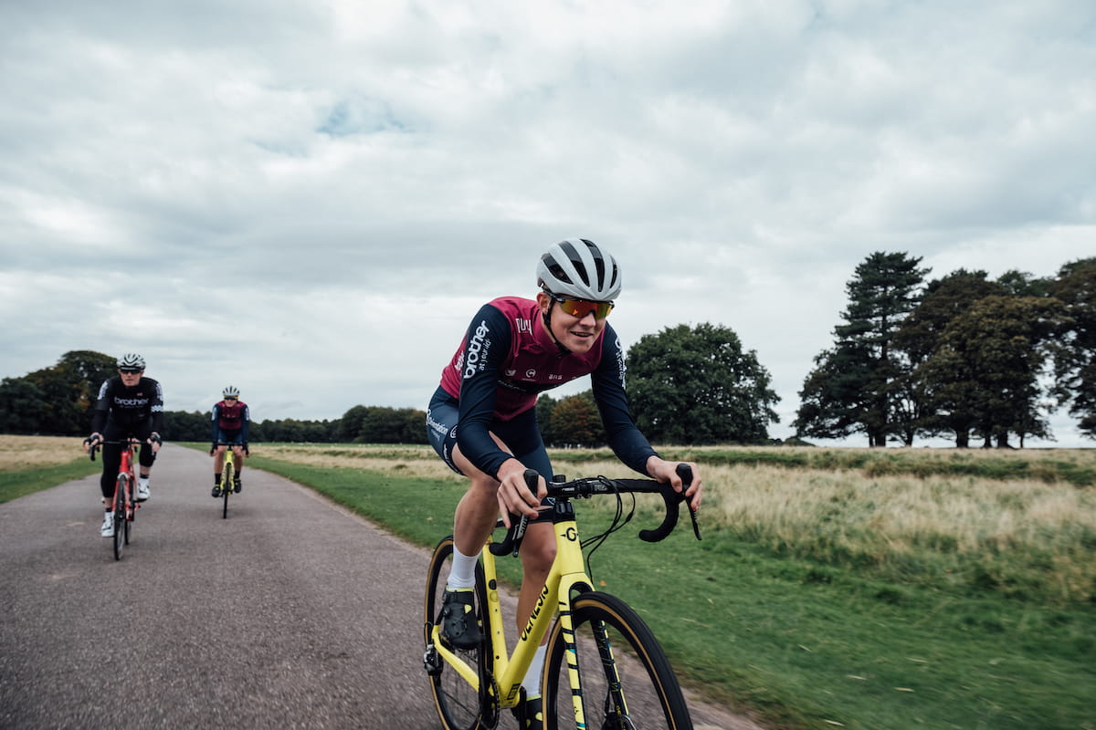 Racing cyclist, purple and blue jersey, blue shorts, white helmet, sunglasses, riding blue and yellow bike with trees and parkland in background