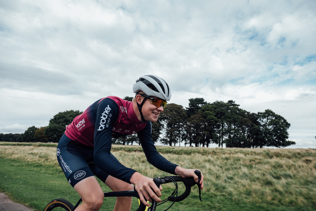 Racing cyclist, purple and blue jersey, blue shorts, white helmet, sunglasses, riding yellow bike, trees and green parkland in background