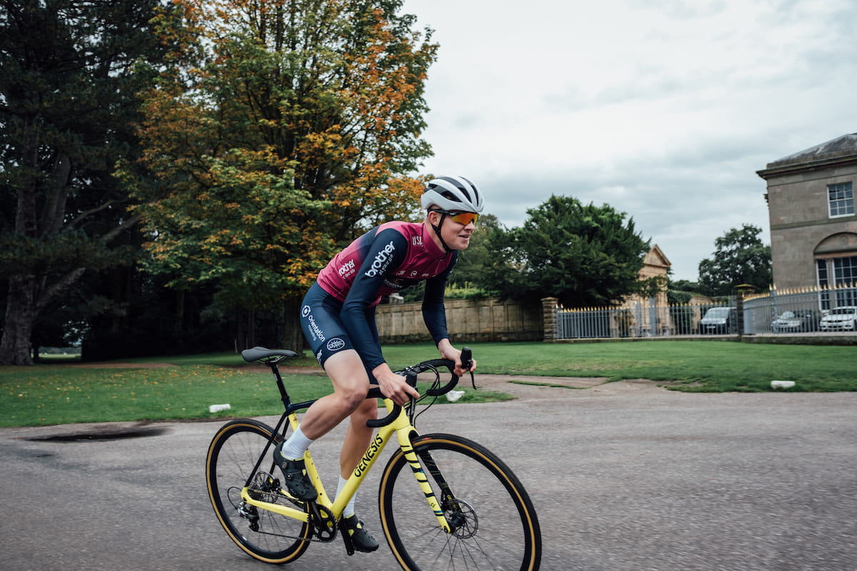 Racing cyclist, purple and blue jersey, blue shorts, white helmet, sunglasses, white socks, black shoes, riding yellow bike past stone house