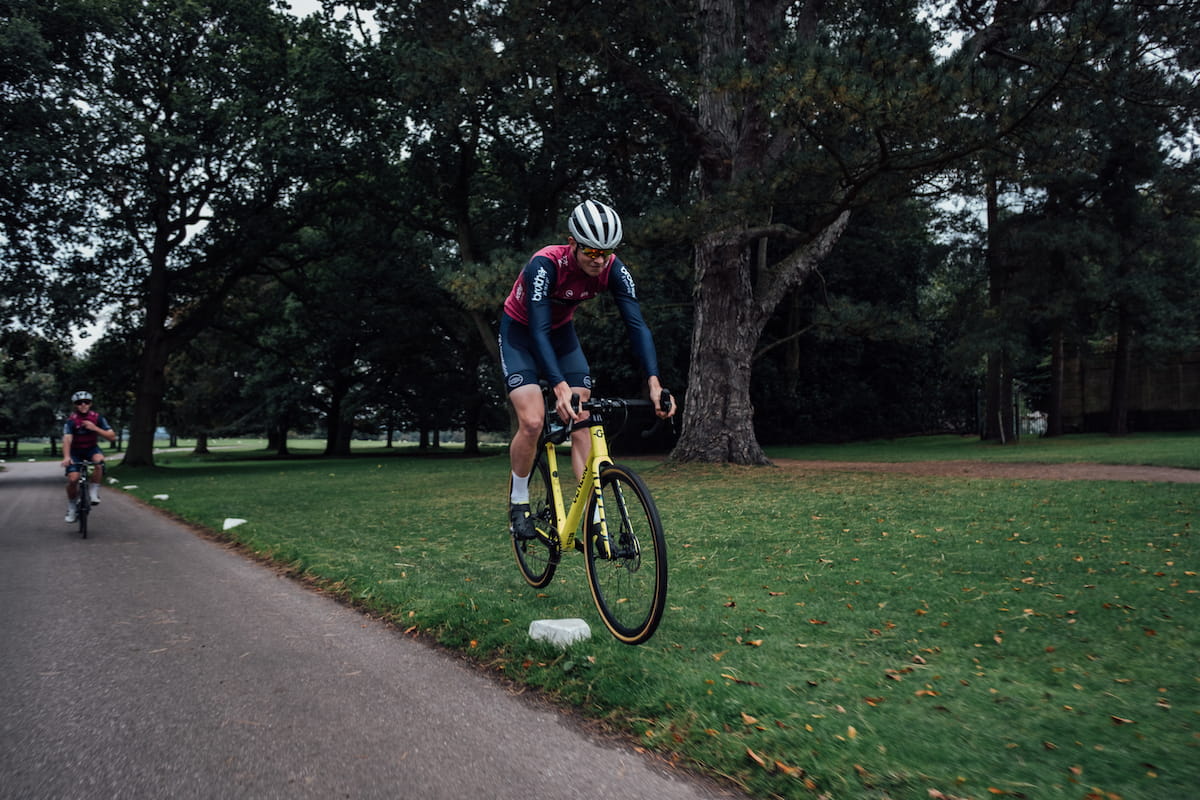 Racing cyclist, purple and blue jersey, blue shorts, white helmet, sunglasses, white socks, black shoes, jumping yellow bike over stone