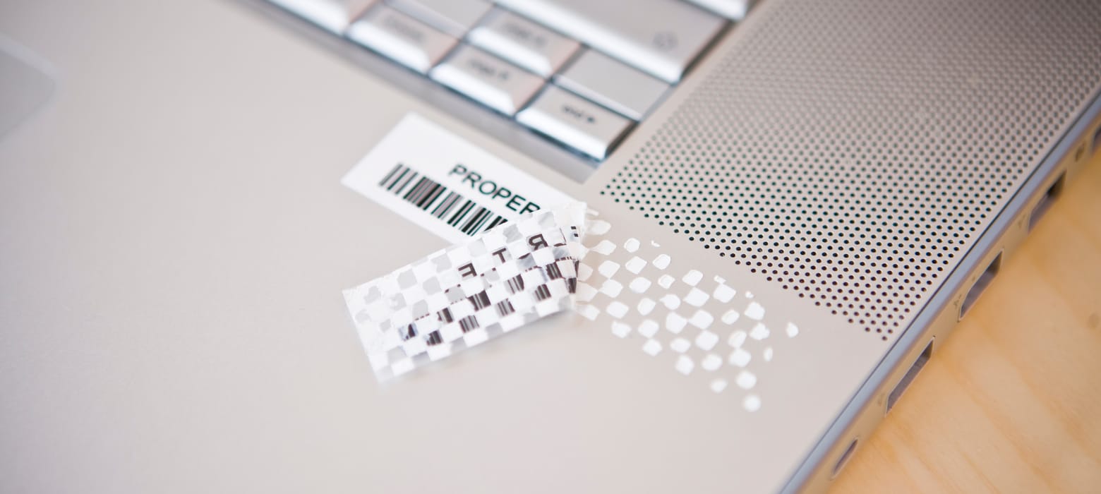 A black on white anti-tamper laminated tape applied to a silver notebook computer has been pulled back to reveal a checkerboard pattern, rendering it unusable