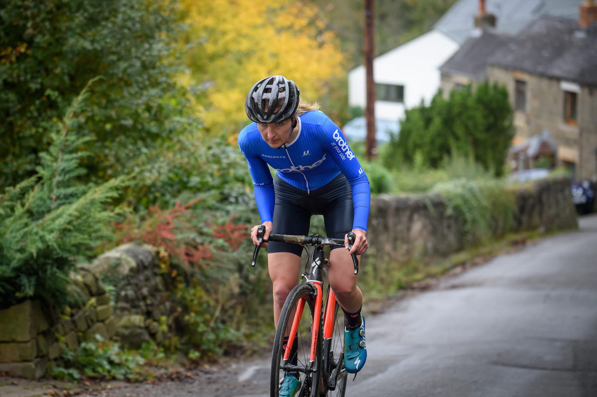 Cyclist Rebecca Richardson riding uphill on a country lane with greenery and cottages is the background