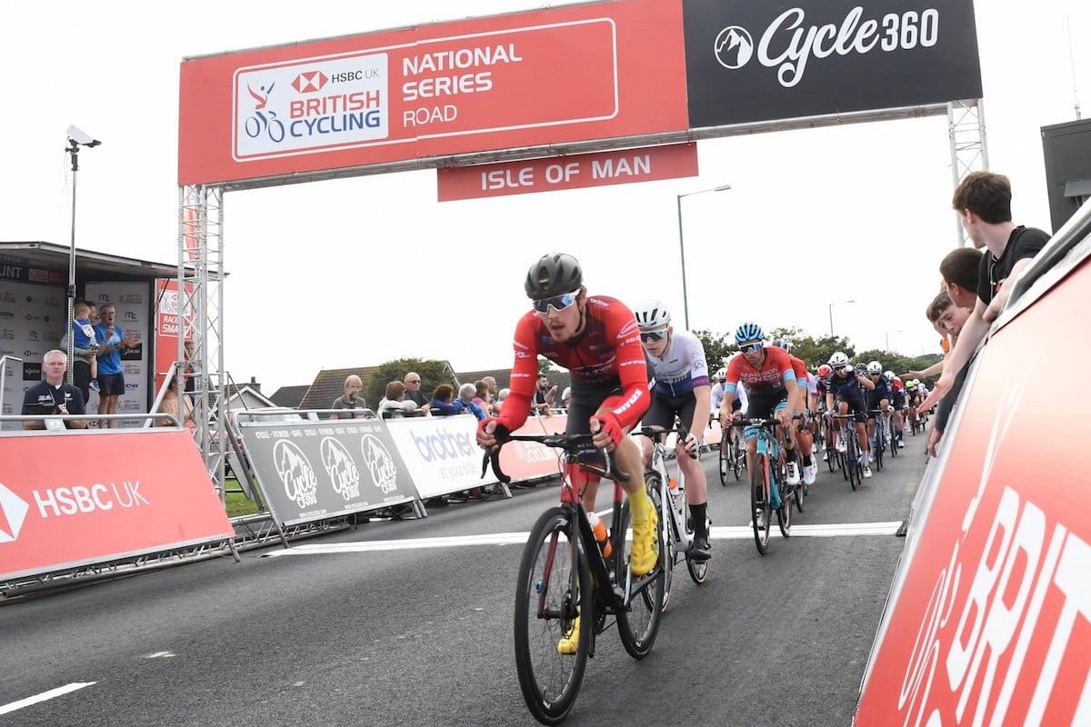 Racing cyclist, male, red jersey, black shorts, yellow shoes, leading line of riders beneath start-finish gentry