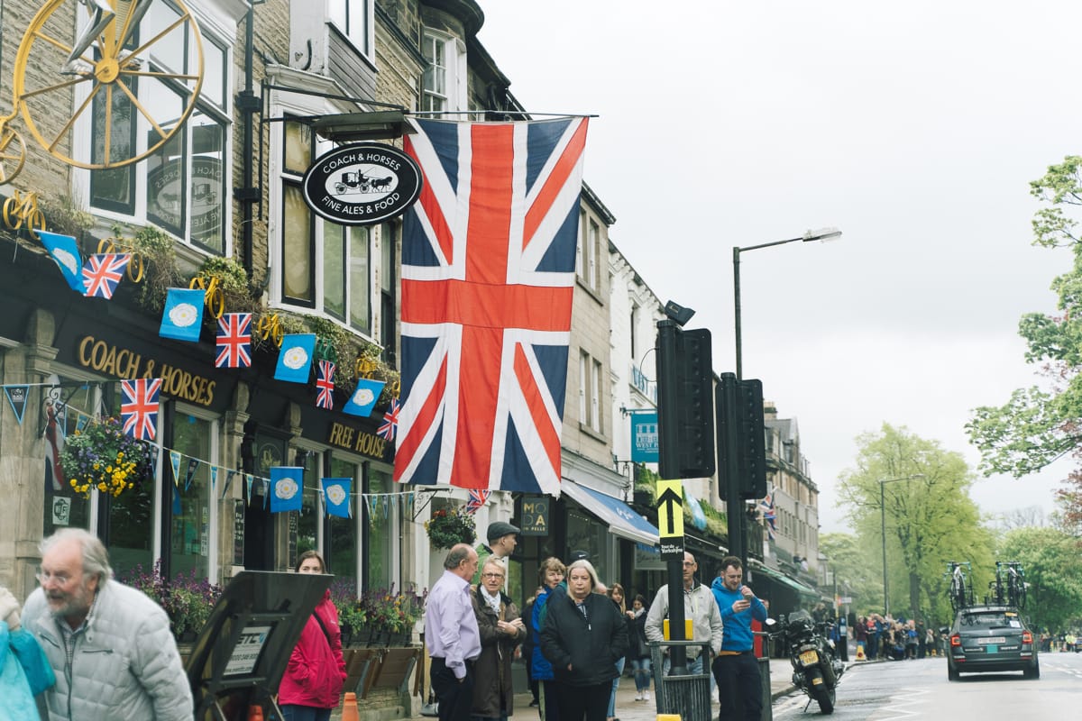 Spectators lining the side of a road in front of a pub adorned with flags