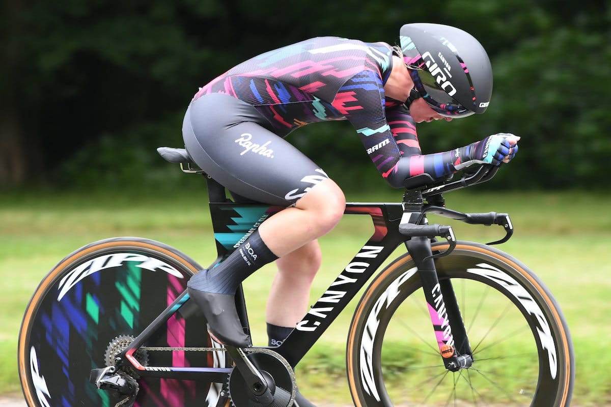 Side view of cyclist Alice Barnes as she has hear head down while riding along a road with greenery in the background