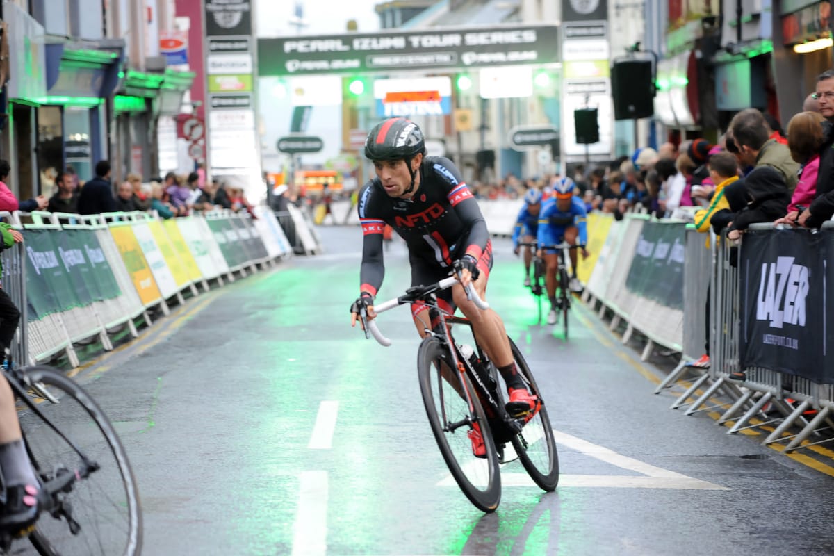 Cyclist Dean Downing taking a bend in the road with spectators behind barriers on either side in a built up area