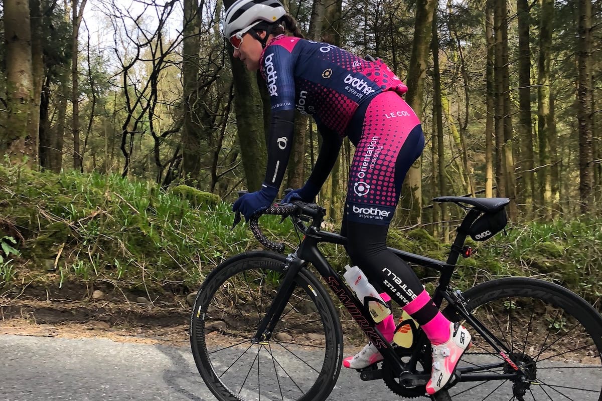 Cyclist Melissa Greaves standing and leaning over the handlebars while riding along a country road with trees and greenery in the background