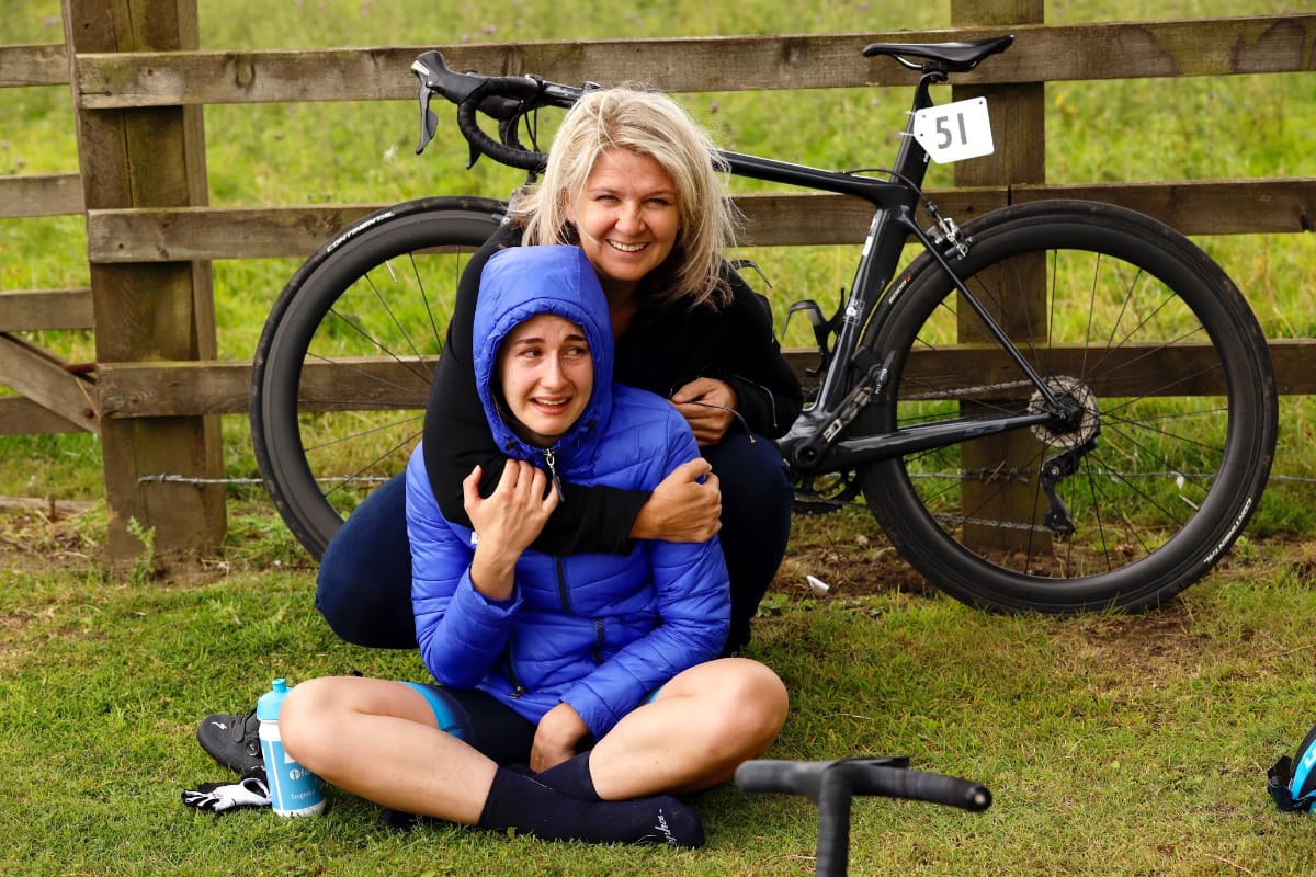 Volunteer VIcky Creer with her arm around team member while kneeling on grass in front of bike which is leaning against fence
