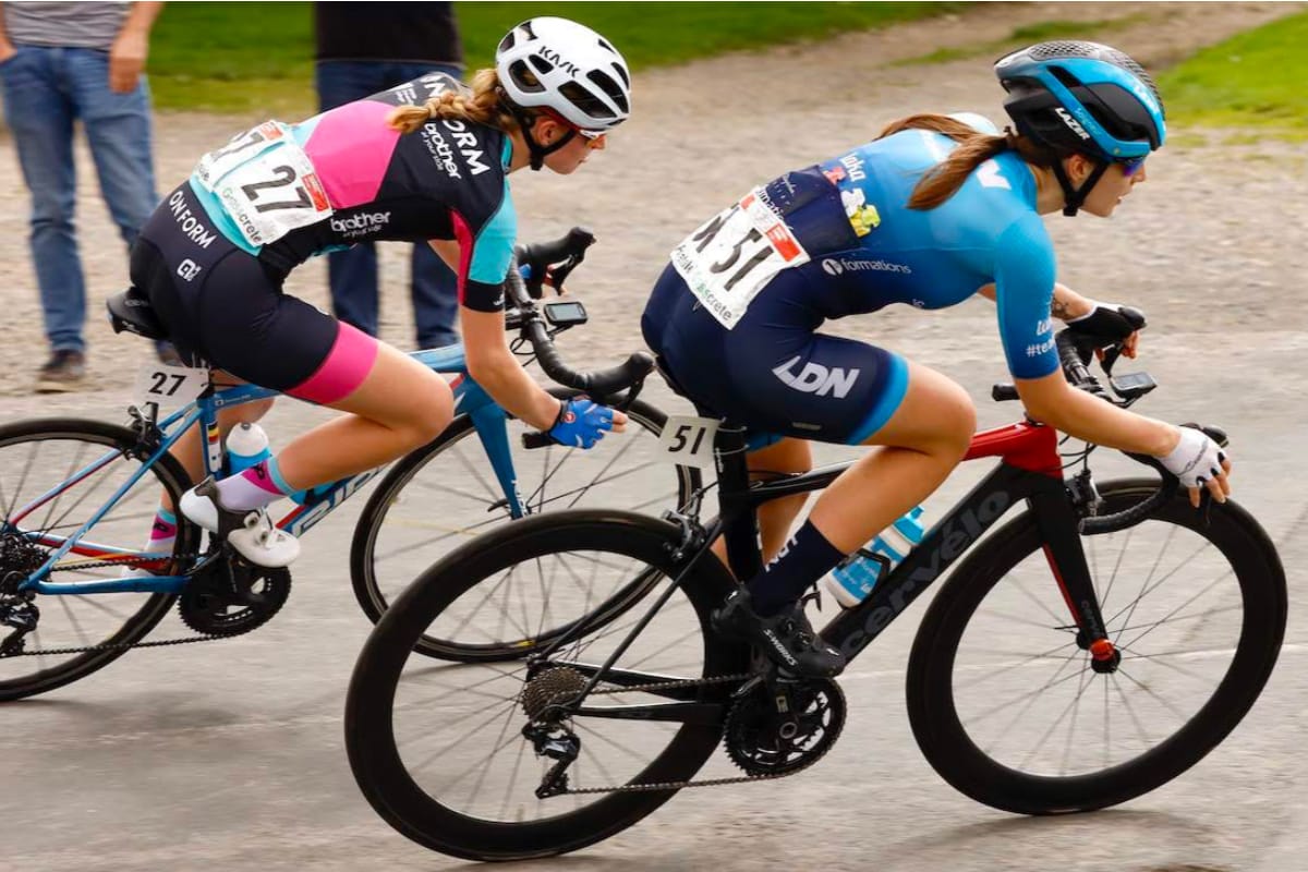Cyclist Fran Cutts leaning into a bend with another cyclist close behind as they pass two spectators in the background
