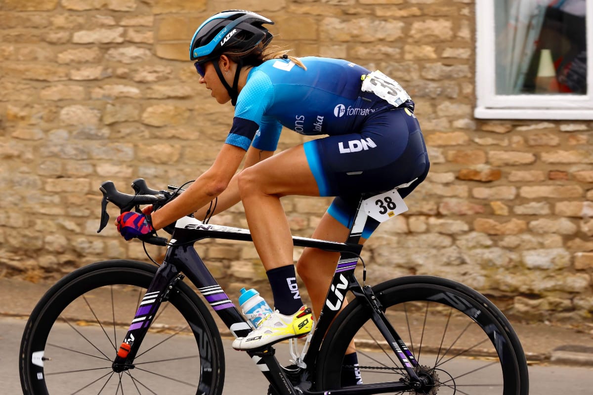 Side view of Cyclist Danni Shrosbree riding along a road with a stone brick wall and window in the background