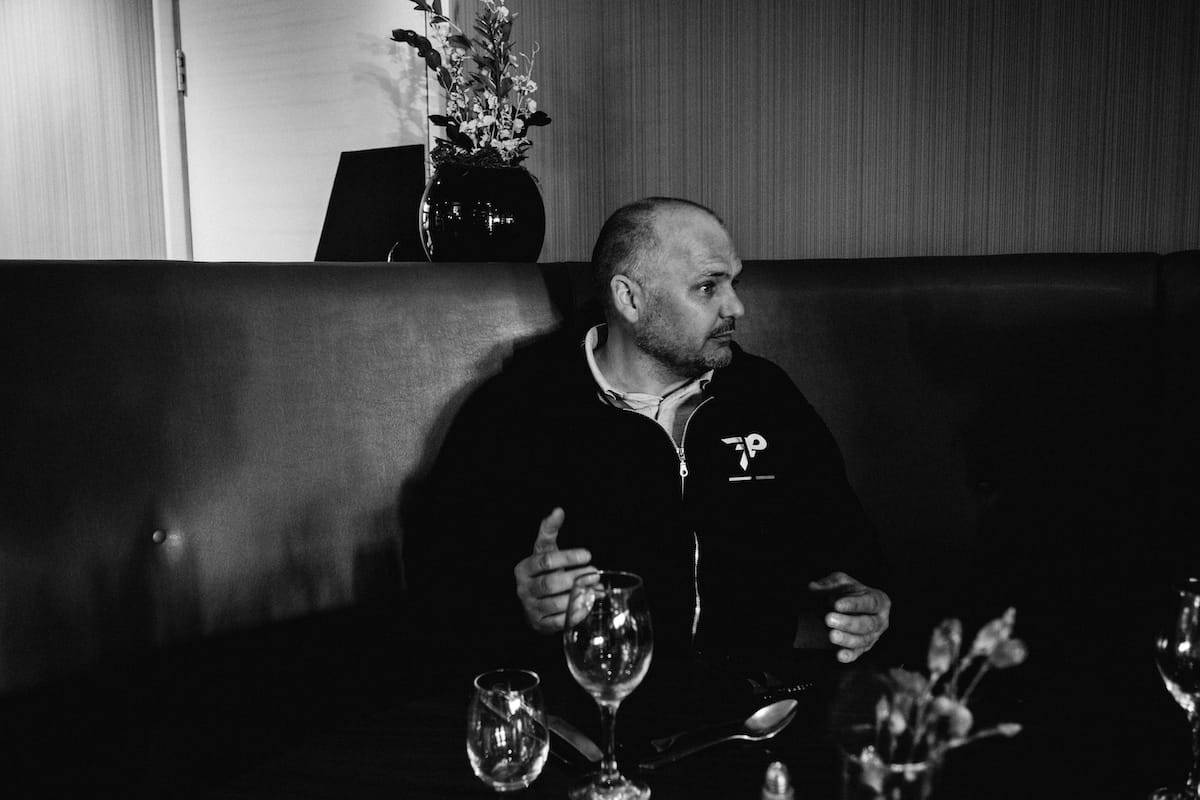 Simon Howes gesturing while talking to someone who is out of shot while sat on a corner sofa with empty wine glasses in the foreground